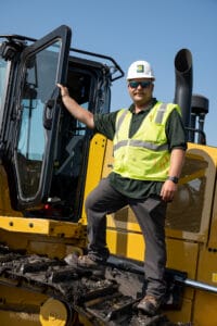 Man in white hard hat and yellow safety vest with sunglasses posing as he opens the door to the cab of a bulldozer.