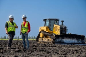 Two men in hard hats and safety vests standing in a dirt field with a bulldozer in the background.