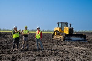 Three men in hard hats and safety vests standing in a field talking with a bulldozer in the background.