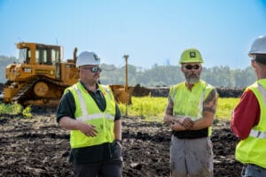 Closeup of three men in hard hats and safety vests standing in a field talking with a bulldozer in the background.