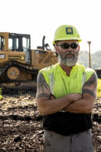 Older gentleman with yellow hard hat, black glasses and safety vest standing with arms crossed in a dirt field with bulldozer in the background.