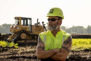 Older gentleman with yellow hard hat, black glasses and safety vest standing with arms crossed and head looking to the side in a dirt field with bulldozer in the background.