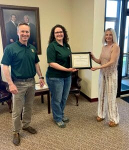 Two women jointly holding up a safety merit award with a man standing to the left in a business lobby.