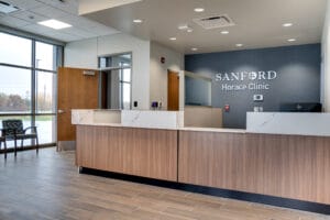 Lobby view looking at receptionists station with blue accent wall with the words "Sanford Horace Clinic" behind and floor to ceiling window wall with waiting chairs to the left.