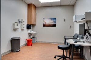 Interior view from doorway looking into patient exam room. Standard desk and swivel chair workstation with sink, garbage cans and basic exam equipment.