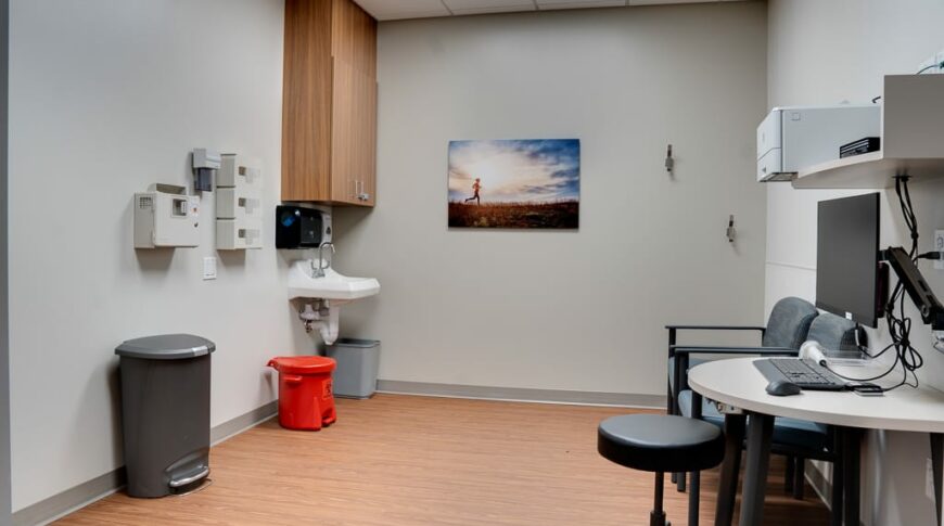 Interior view from doorway looking into patient exam room. Standard desk and swivel chair workstation with sink, garbage cans and basic exam equipment.