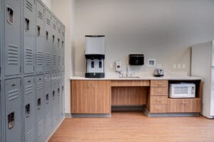 Breakroom area with gray lockers lining the lefthand side wall and a convenience station to the back wall with lower wood cabinetry and sink and coffee machine atop with built-in microwave and fridge.