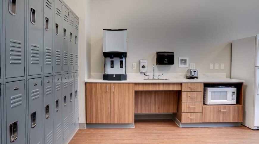 Breakroom area with gray lockers lining the lefthand side wall and a convenience station to the back wall with lower wood cabinetry and sink and coffee machine atop with built-in microwave and fridge.