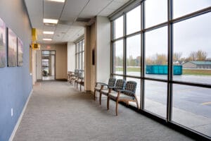 Waiting hallway with wall of floor to ceiling windows to the right with waiting chairs and blue accent wall with pictures to the left.