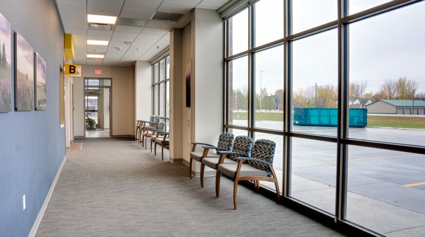 Waiting hallway with wall of floor to ceiling windows to the right with waiting chairs and blue accent wall with pictures to the left.