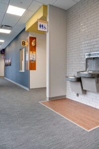 Waiting hallway with two water fountains with white subway tile accent wall behind to the right. Bathroom sign overhead and passthrough reception window to the far wall at the Sanford Horace Clinic.