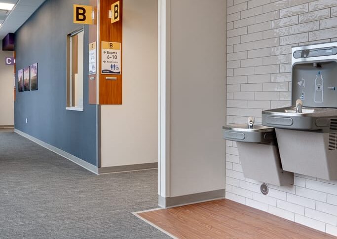 Waiting hallway with two water fountains with white subway tile accent wall behind to the right. Bathroom sign overhead and passthrough reception window to the far wall.