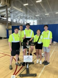 Five people in safety green shirts and shorts standing around an exercise bike for a charity race event.