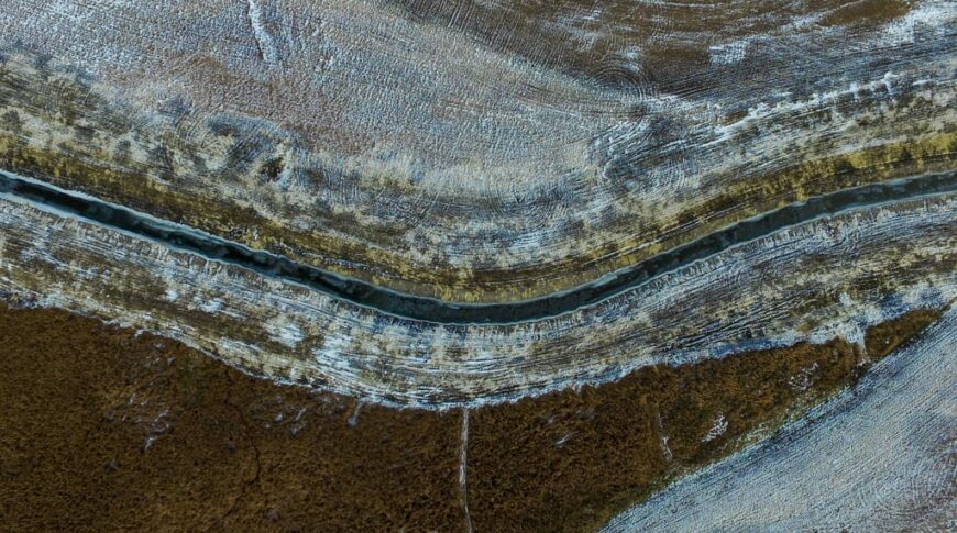 Overhead view looking down at a bend in a county drainage ditch filled with blue water running across pasture land.