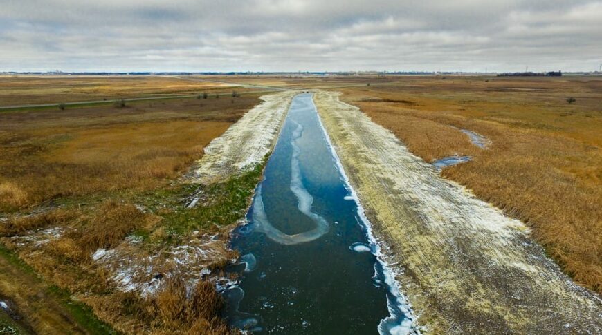 Aerial view of a straight drainage ditch that has recently been excavated with dark blue water running throughout during the fall season.