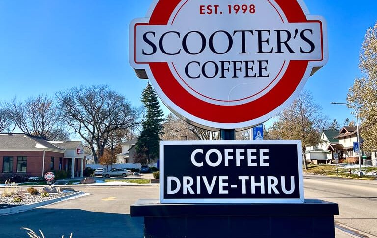 Coffee shop sign at their parking lot entrance noting that they are a coffee drive-thru location. Some rocks and shrubbery underneath for decoation.