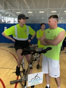 Young man with black baseball hat, yellow safety shirt and black pants riding on an exercise bike while talking to another man in yellow safety shirt and shorts.