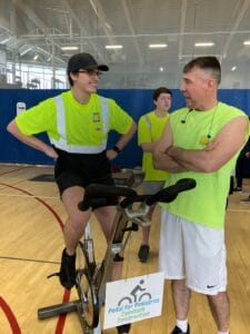 Young man with black baseball hat, yellow safety shirt and black pants riding on an exercise bike while talking to another man in yellow safety shirt and shorts.