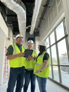 Three people in hard hats and safety vests laughing while standing in a construction jobsite indoors.