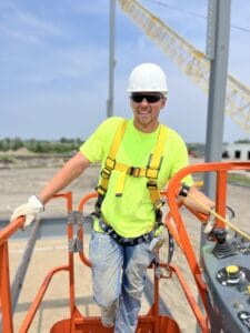 Young man in white hard hat and safety clothes harnessed to a lift truck overlooking a construction jobsite.