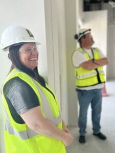 Woman in hard hat and safety vest smiling with man blurred in background also in hard hat and safety vest on the interior of an unfinished jobsite.