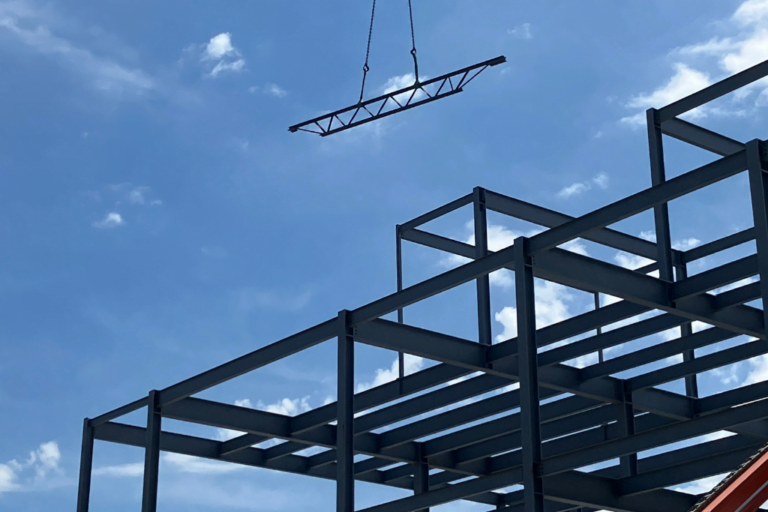 Building metal beams with a metal beam hovering in place being put in place by a crane.