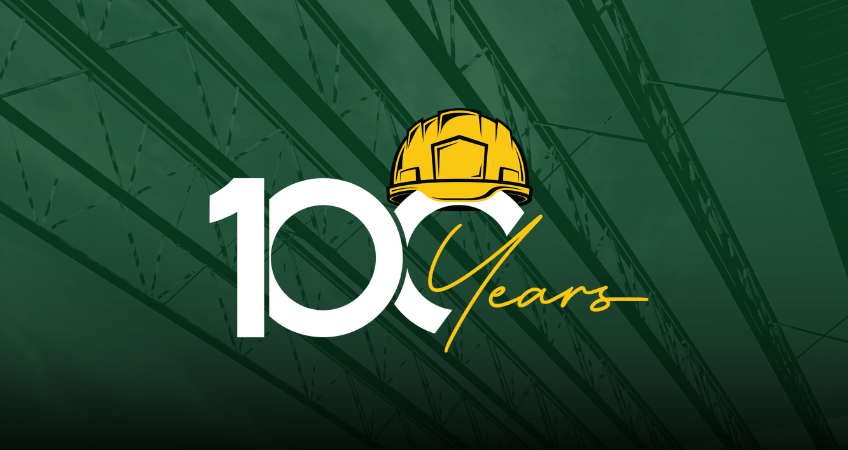 100-year logo with yellow hard hat icon and background image in green overlay of metal beams at a construction jobsite. Our 100-year theme is Building our Communities since 1924, one vision at a time.