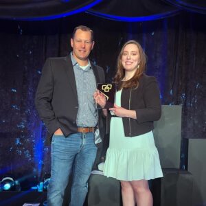 Man in jeans, button-up chequed shirt and black jacket standing next to woman in white dress and black jacket holding a plaque with a dark blue lighted background.