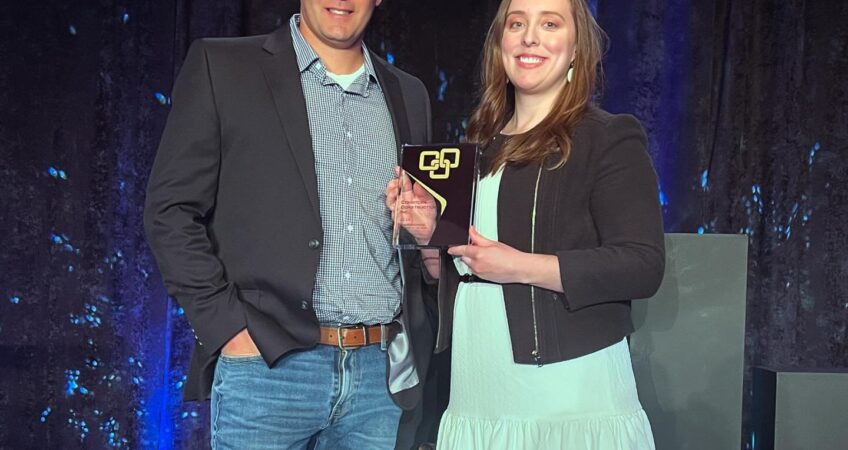 Man in jeans, button-up chequed shirt and black jacket standing next to woman in white dress and black jacket holding a plaque with a dark blue lighted background.