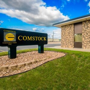 Brown brick Comstock Construction office building with sign in the foreground on grassy knoll with bright blue and clouds sky in background in Wahpeton, ND