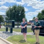 Michael Comstock stands on grassy boulevard next to Bradley Aune from the FMWF Chamber of Commerce, giving a talk about the history of Comstock Construction.