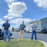 Comstock Construction's rebar cutting celebration with three men holding a green piece of rebar with the middle one also holding a cutting tool. Background of the Comstock Construction building in Fargo with blue sky and clouds.