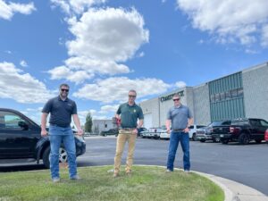 Comstock Construction's rebar cutting celebration with three men holding a green piece of rebar with the middle one also holding a cutting tool. Background of the Comstock Construction building in Fargo with blue sky and clouds.