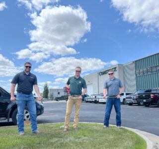 Comstock Construction's rebar cutting celebration with three men holding a green piece of rebar with the middle one also holding a cutting tool. Background of the Comstock Construction building in Fargo with blue sky and clouds.