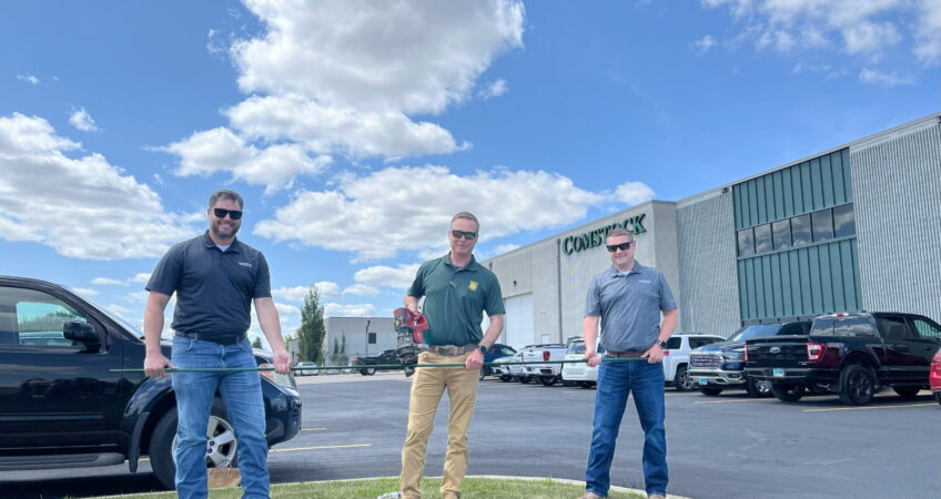 Comstock Construction's rebar cutting celebration with three men holding a green piece of rebar with the middle one also holding a cutting tool. Background of the Comstock Construction building in Fargo with blue sky and clouds.