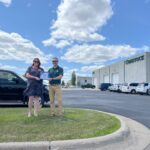 Man and woman stand next to each other on grassy knoll, each holding a plaque. Parking lot and blue sky with clouds in the background.