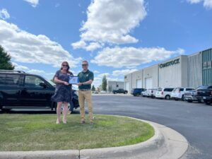 Man and woman stand next to each other on grassy knoll, each holding a plaque. Parking lot and blue sky with clouds in the background.