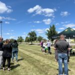 About ten people standing on grass with tent in background and blue sky with clouds.