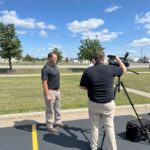 Man being interviewed by reporter from WDAY with grass, trees, and blue sky with clouds in the background.