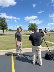 Man being interviewed by reporter from WDAY with grass, trees, and blue sky with clouds in the background.