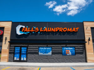 Exterior of black building with the words "Falls Laundromat" in orange letters. Behind is a blue sky with white clouds.