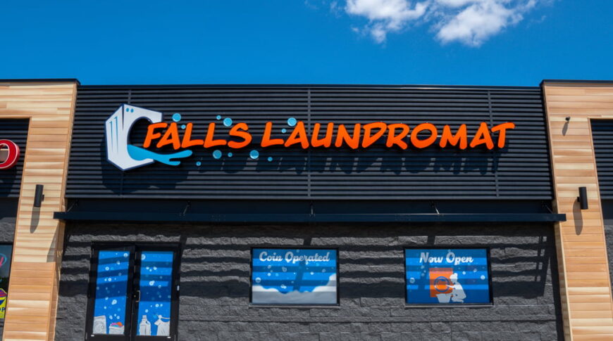Exterior of black building with the words "Falls Laundromat" in orange letters. Behind is a blue sky with white clouds.