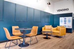 Lounge area at laundromat with faux leather chairs around a table and more chairs behind. Machine detergent vending machine on back wall.