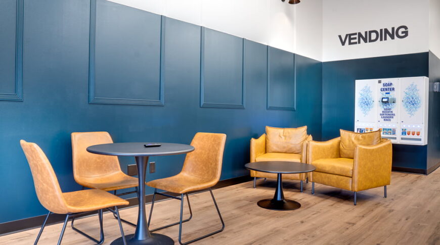 Lounge area at laundromat with faux leather chairs around a table and more chairs behind. Machine detergent vending machine on back wall.