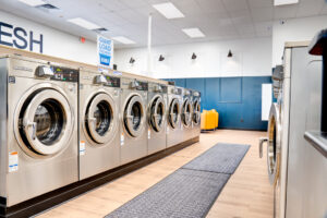 Row of silver washing machines at a laundromat with white oak flooring underneath and lounge area in background.