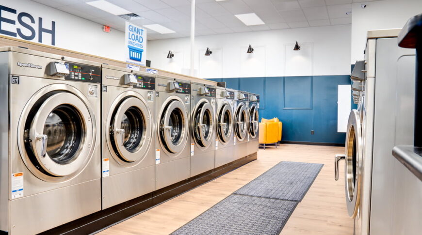 Row of silver washing machines at a laundromat with white oak flooring underneath and lounge area in background.