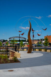 Tall rust-colored tower sculptures with orange butterflies hanging on a pendulum around it. Bright blue sky with clouds in the background.