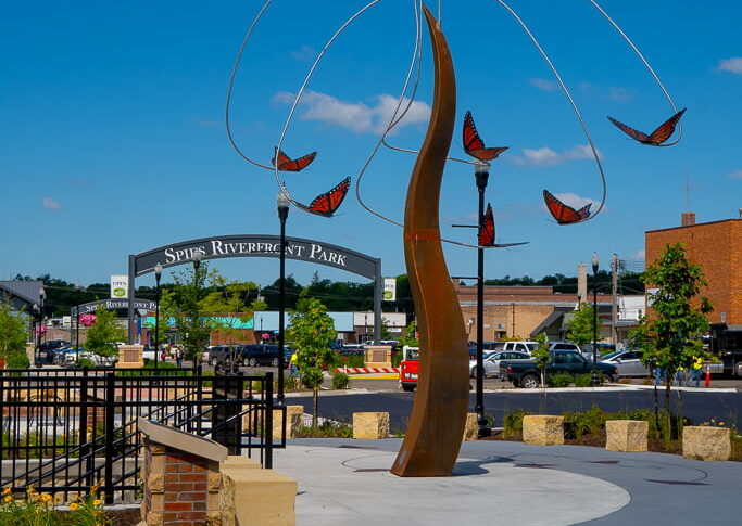 Tall rust-colored tower sculptures with orange butterflies hanging on a pendulum around it. Bright blue sky with clouds in the background.