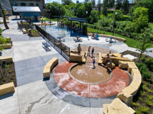 Aerial view of a circular wading pool with concrete rock seating formations around it. Water trickling down to small waterfall with splash pad below.