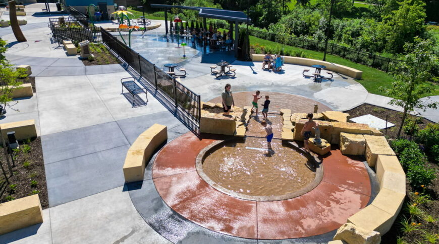 Aerial view of a circular wading pool with concrete rock seating formations around it. Water trickling down to small waterfall with splash pad below.